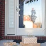 A stack of beautifully wrapped gifts sits on a doorstep with a reflective door and decorative wreath.